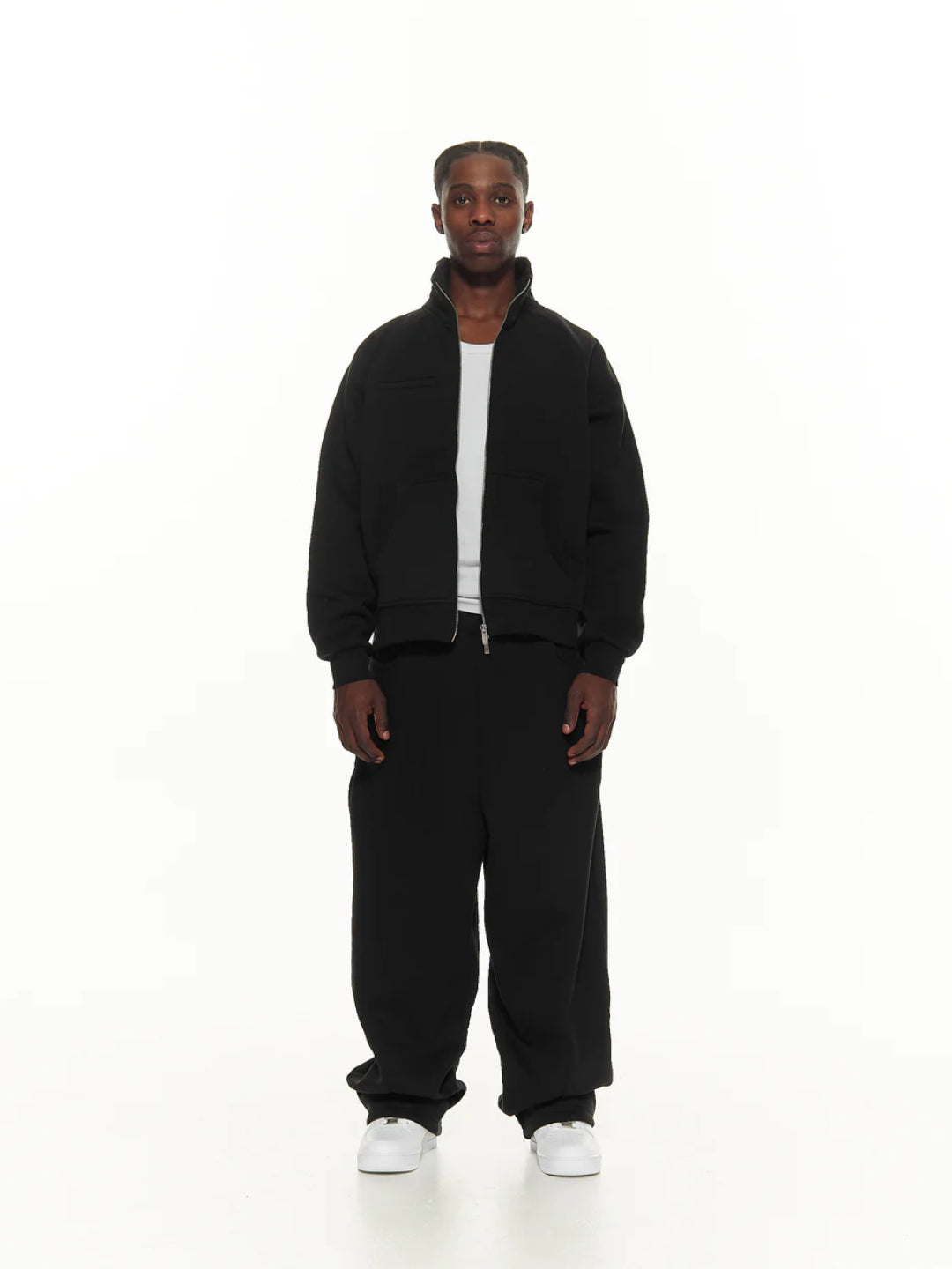 A man standing against a white background, wearing a black tracksuit with a hoodie and pants.