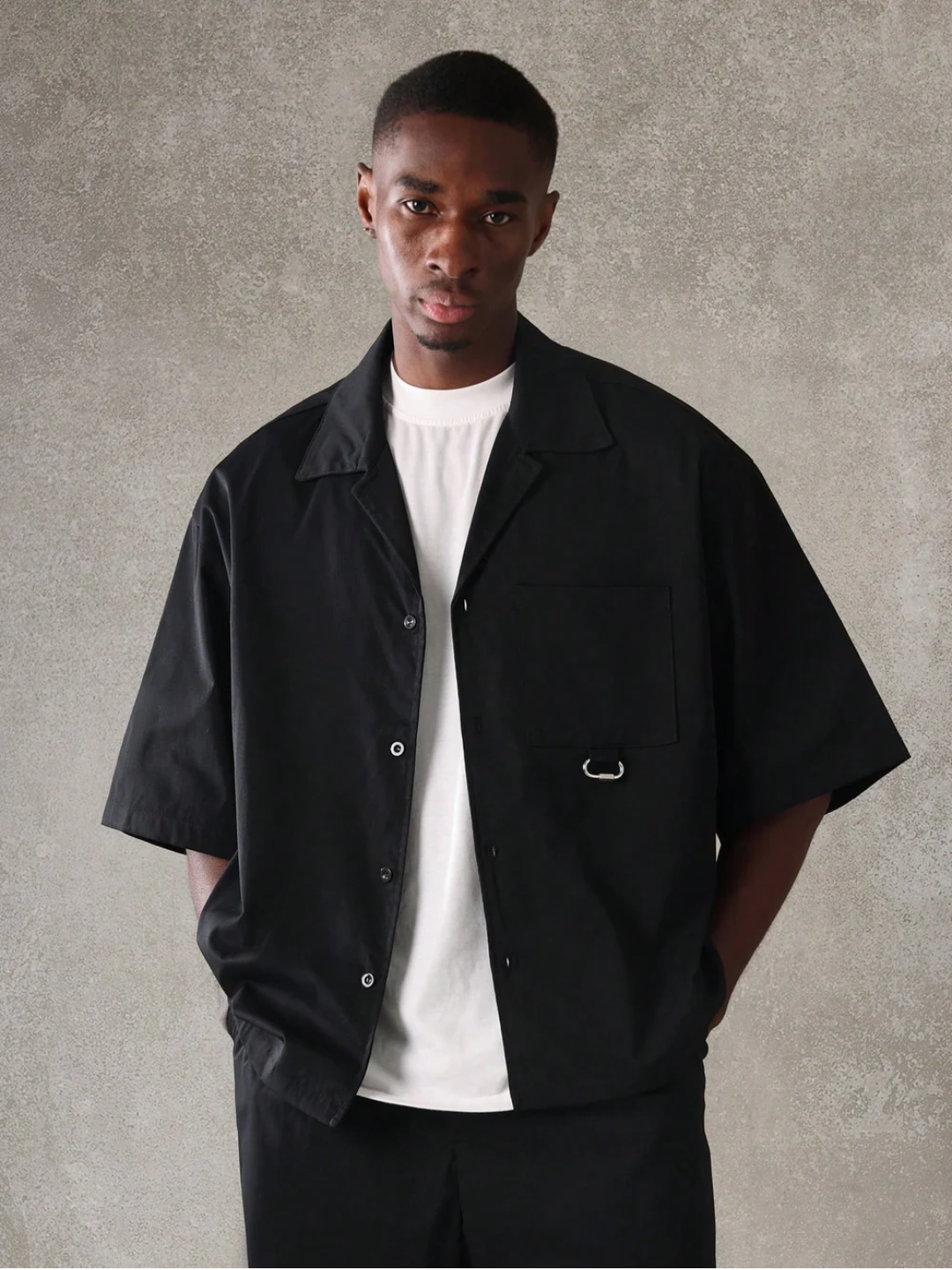 A male model wearing a Black crop fit shirt with a classic collar and drop shoulder sleeves, paired with beige loose fit pants.