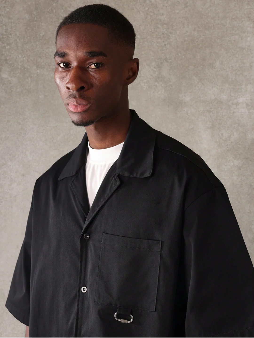 A male model wearing a Black crop fit shirt with a classic collar and drop shoulder sleeves, paired with beige loose fit pants.