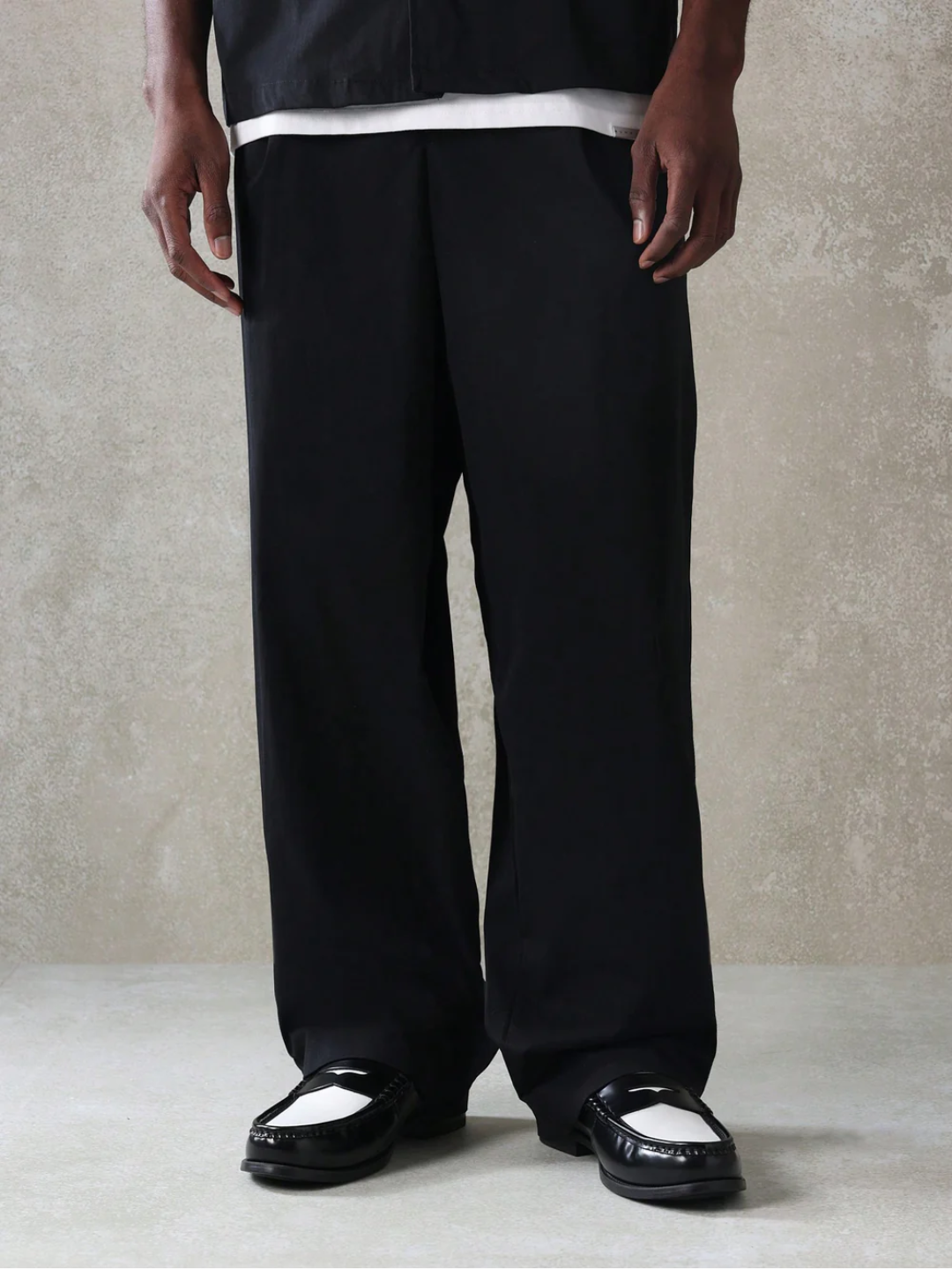 A male model wearing a Black crop fit shirt with a classic collar and drop shoulder sleeves, paired with beige loose fit pants.