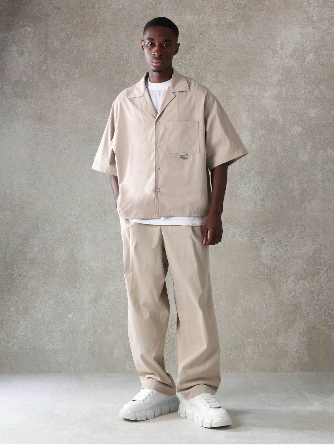 A male model wearing a Beige crop fit shirt with a classic collar and drop shoulder sleeves, paired with beige loose fit pants.
