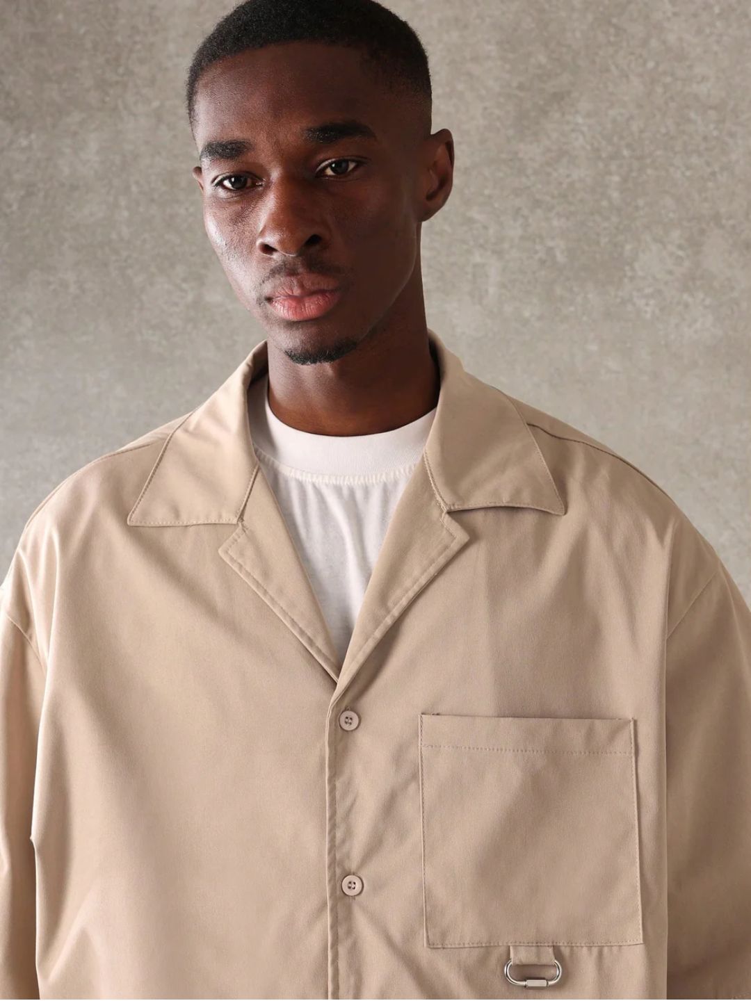 A male model wearing a Beige crop fit shirt with a classic collar and drop shoulder sleeves, paired with beige loose fit pants.