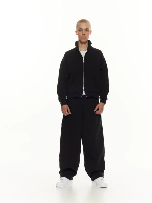A male model wearing a black track jacket with double zippers and a white graphic on the sleeve, standing against a white background.