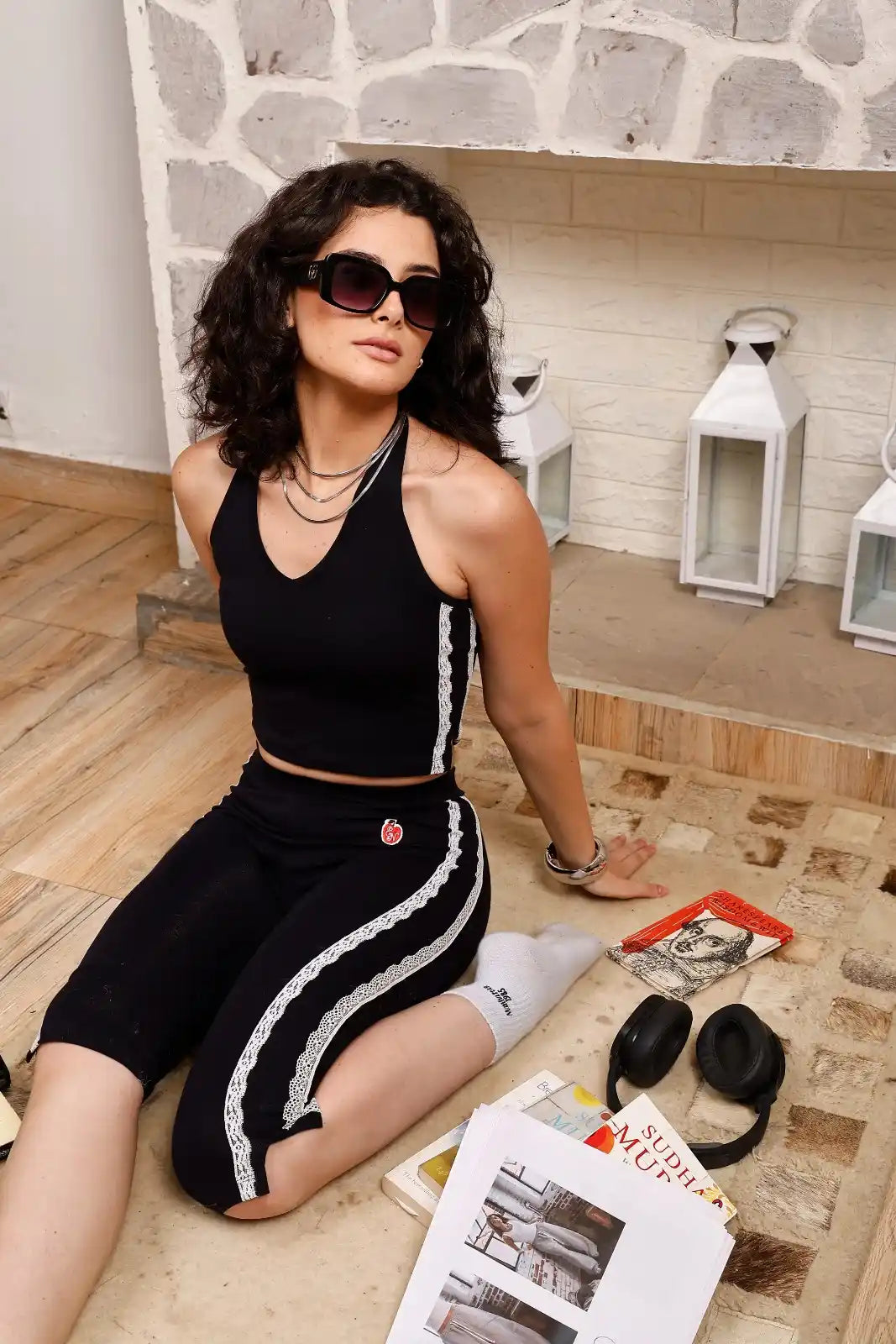 Woman in black outfit sitting on a wooden floor with books and a hat.