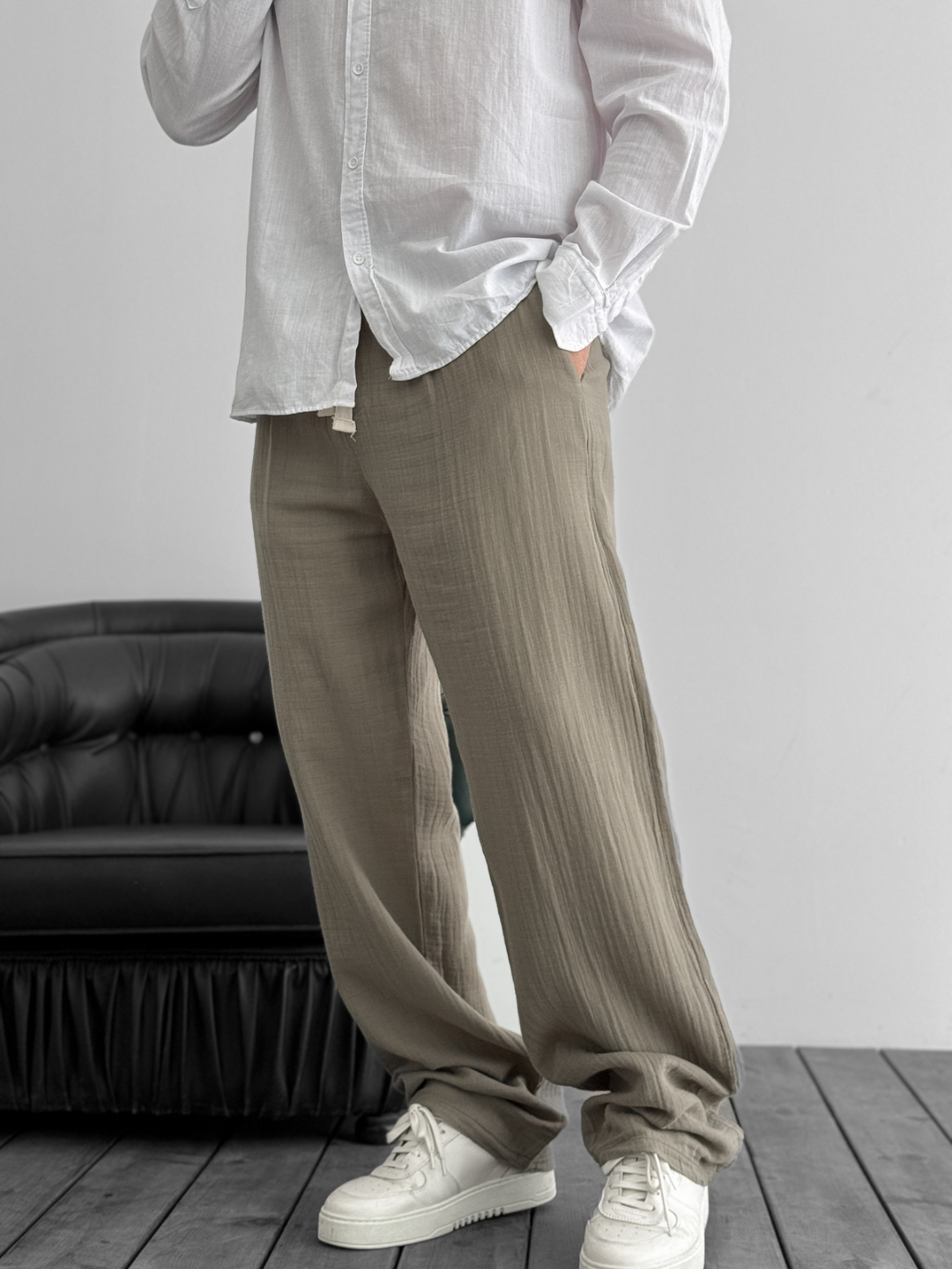 A person standing against a white background, wearing wide-leg gray gauze pants and a white shirt with the sleeves rolled up to the elbows.