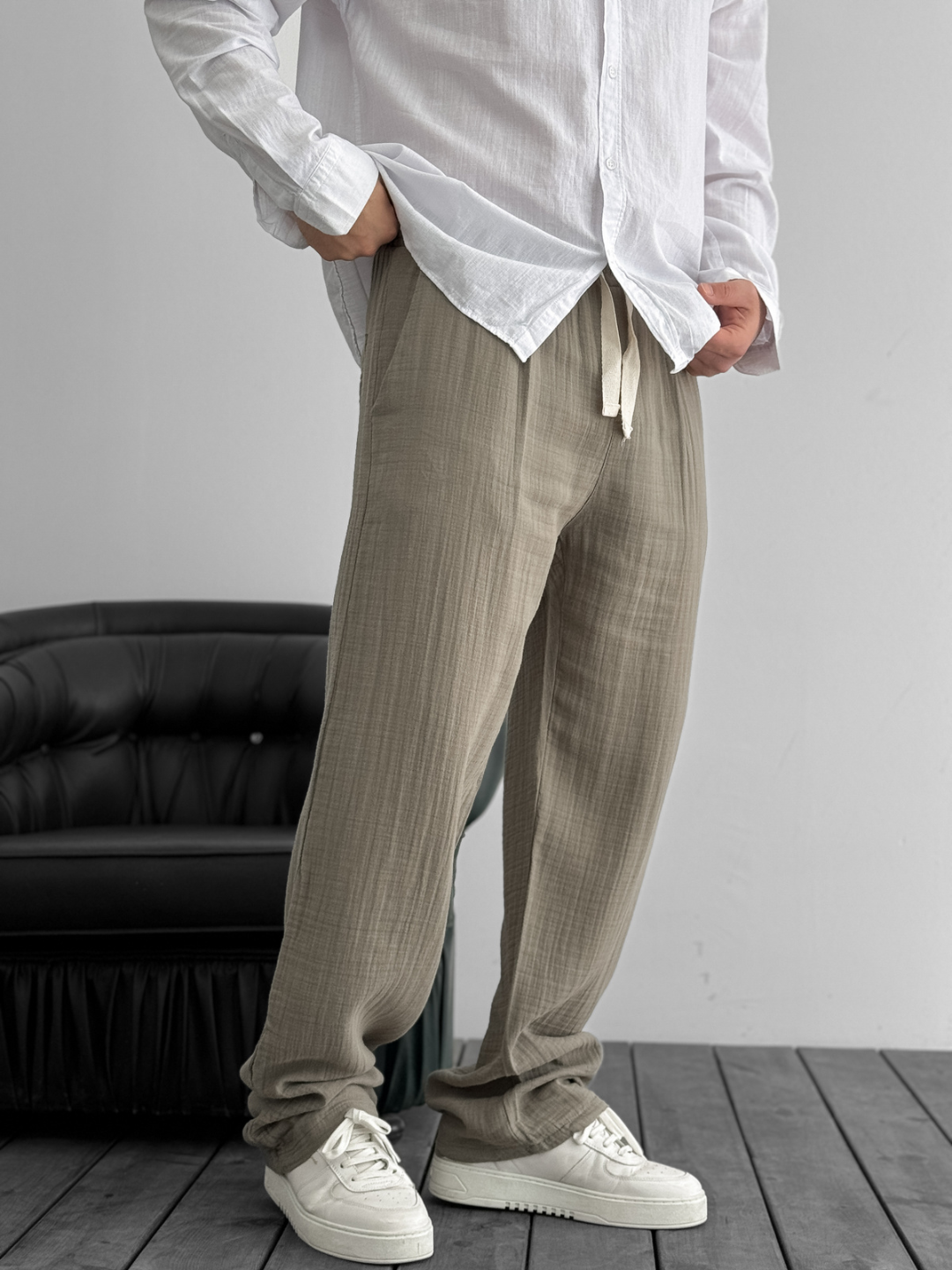 A person standing against a white background, wearing wide-leg gray gauze pants and a white shirt with the sleeves rolled up to the elbows.