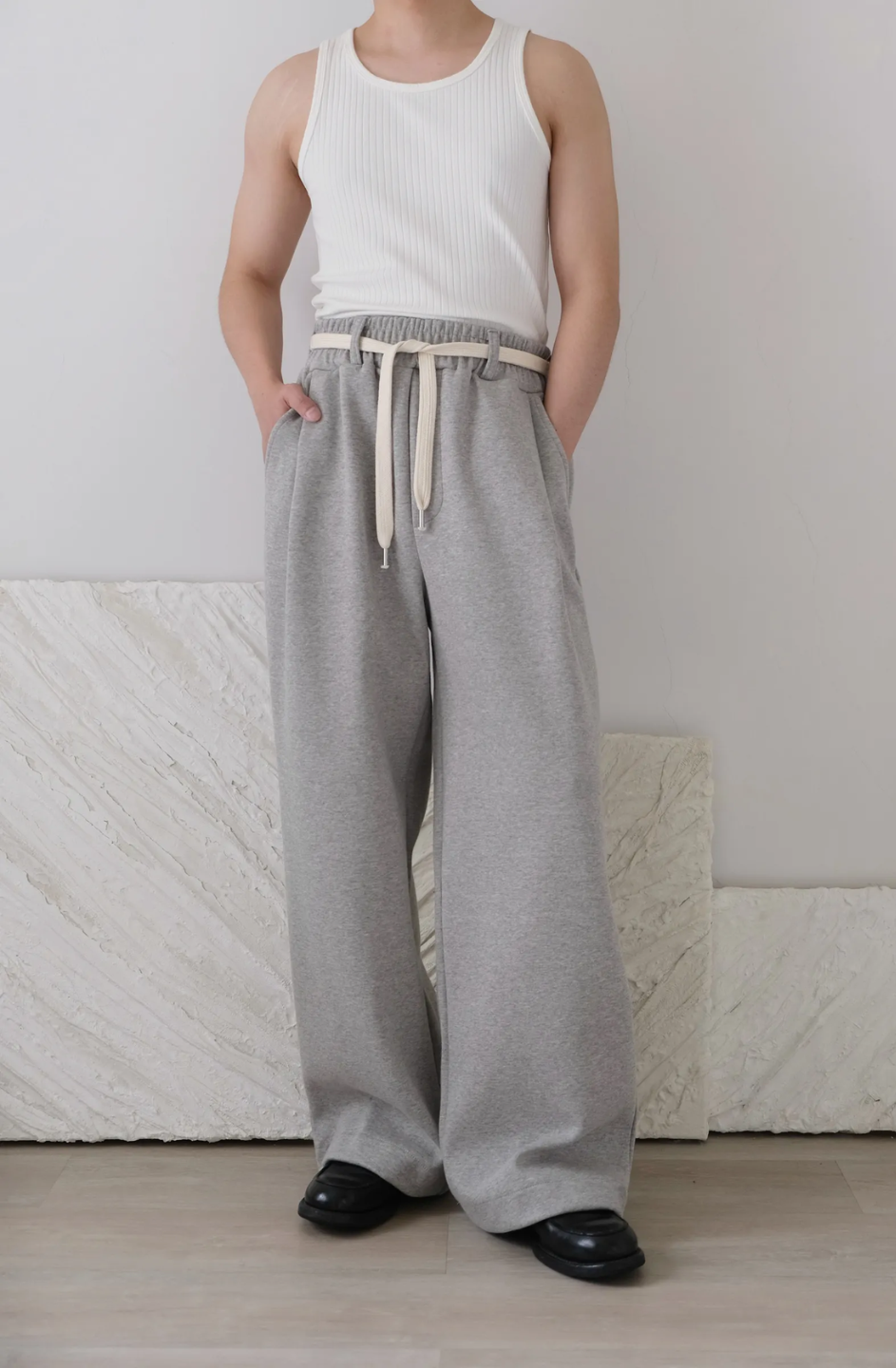 A person standing in a room wearing wide leg gray pants with a cotton belt, a white top, and black shoes.