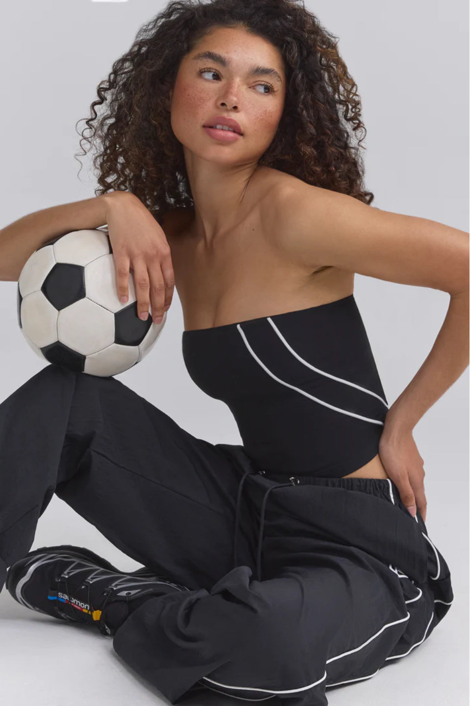 A woman sitting on the floor wearing a black bandeau top and black pants, holding a soccer ball.