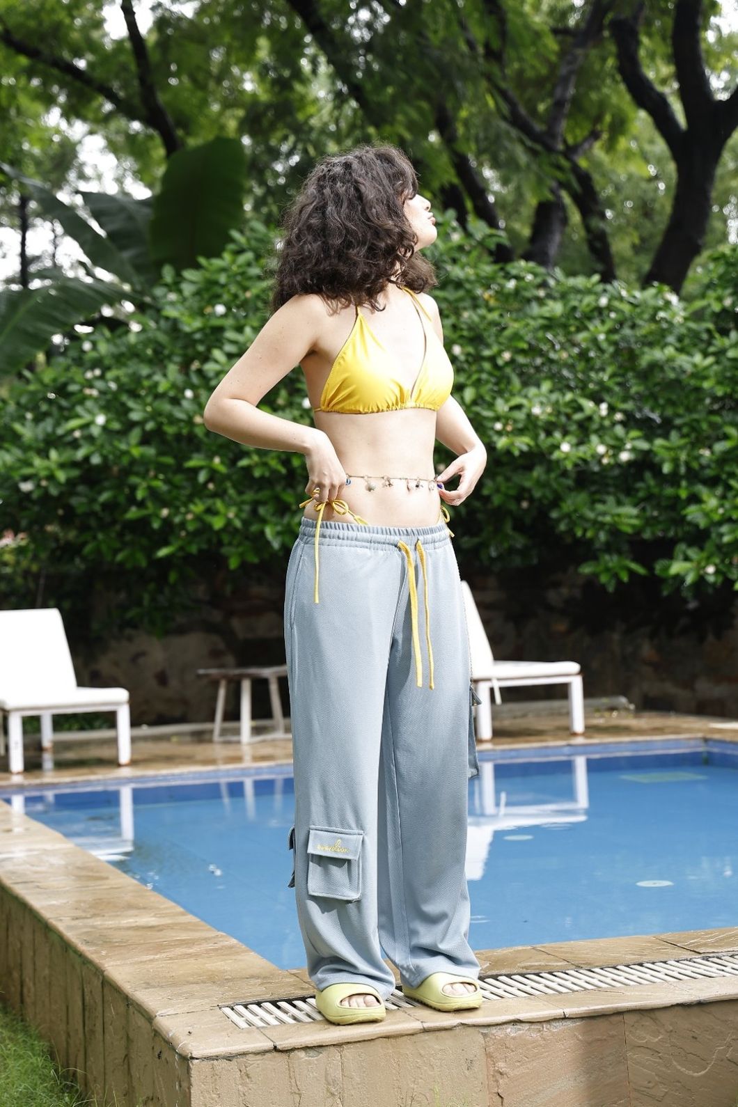 Woman in yellow bikini top and light gray pants standing by a pool with greenery in the background