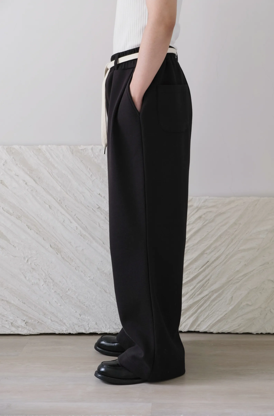 Person wearing black pants and black shoes against a white wall.