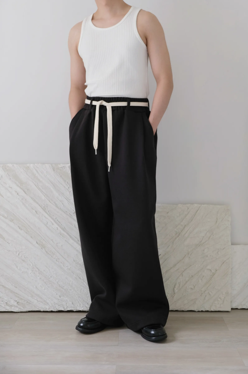 Person wearing a white sleeveless top and black wide-leg pants with a belt, standing against a plain background.