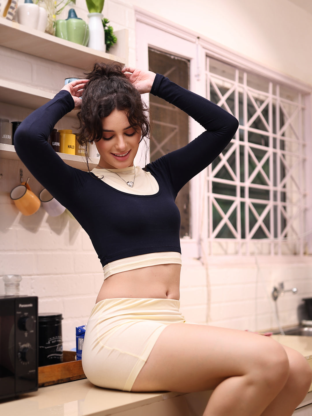 Woman sitting on a kitchen counter wearing a black long-sleeve top and beige skirt.