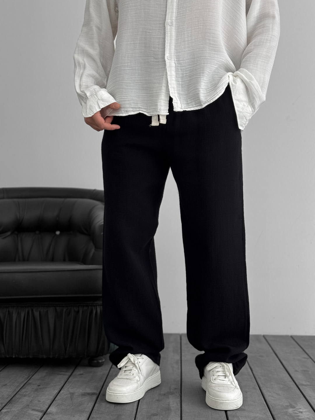 A person standing against a white background, wearing wide-leg gray gauze pants and a white shirt with the sleeves rolled up to the elbows.