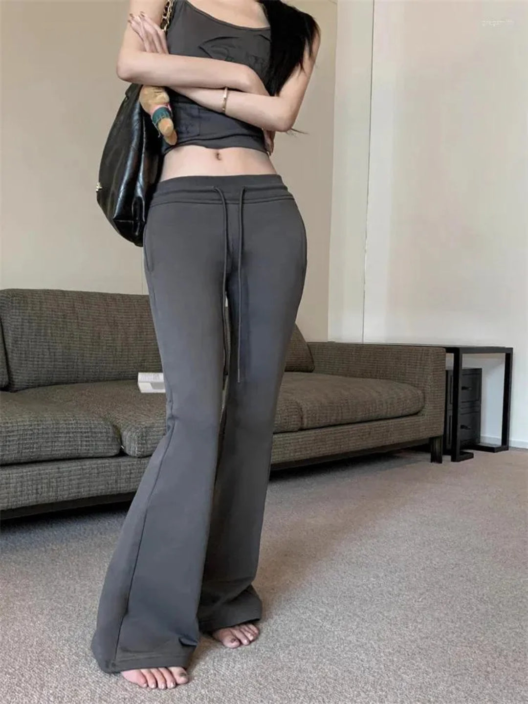 A woman standing in a living room wearing dark grey flared sweatpants with an elasticized waistband and drawstrings, paired with a grey top and black sandals.