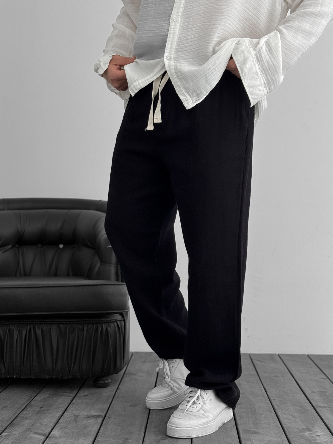 A person standing against a white background, wearing wide-leg black gauze pants and a white shirt with the sleeves rolled up to the elbows.