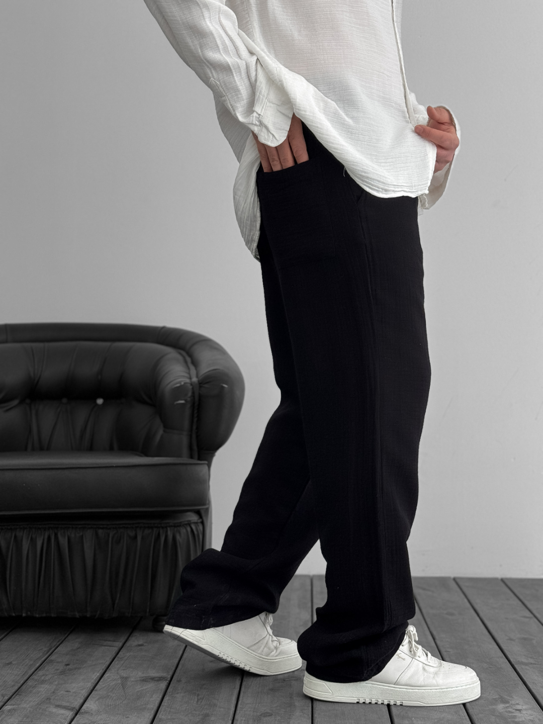 A person standing against a white background, wearing wide-leg black gauze pants and a white shirt with the sleeves rolled up to the elbows.