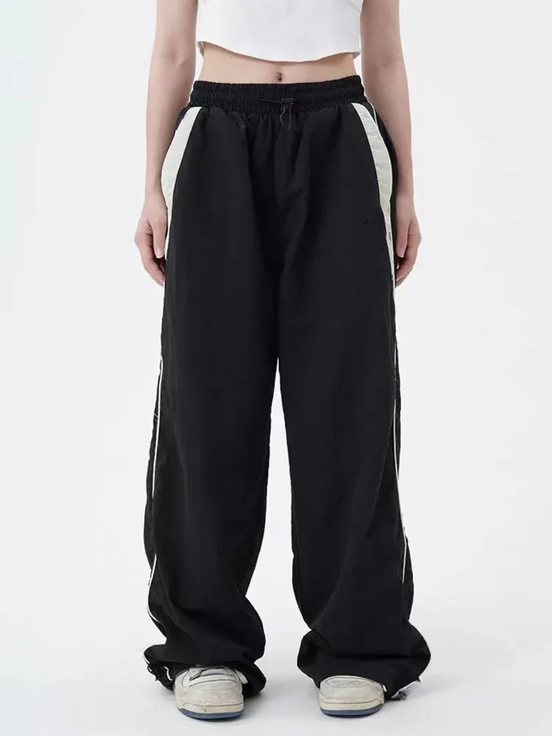 A pair of black track pants with contrasting white piping on the sides, worn by a person standing against a white background.