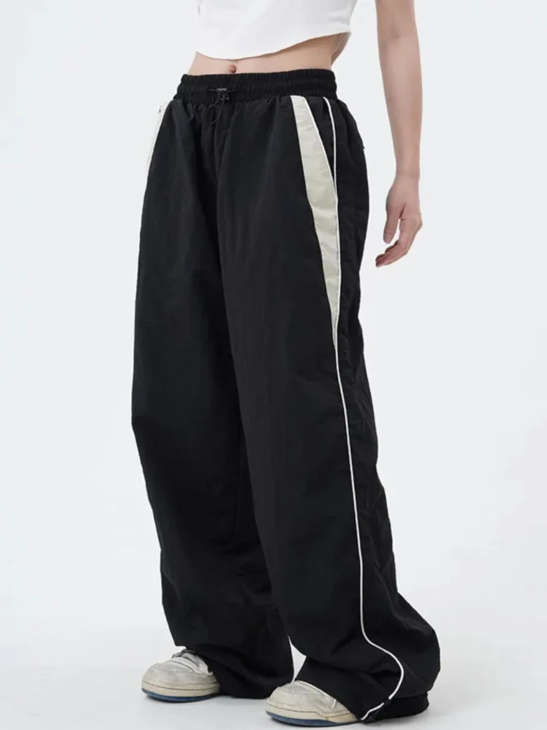 A pair of black track pants with contrasting white piping on the sides, worn by a person standing against a white background.