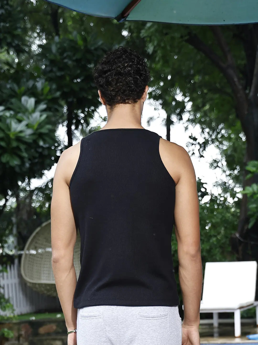 Person wearing a black tank top standing outdoors with trees and a chair in the background