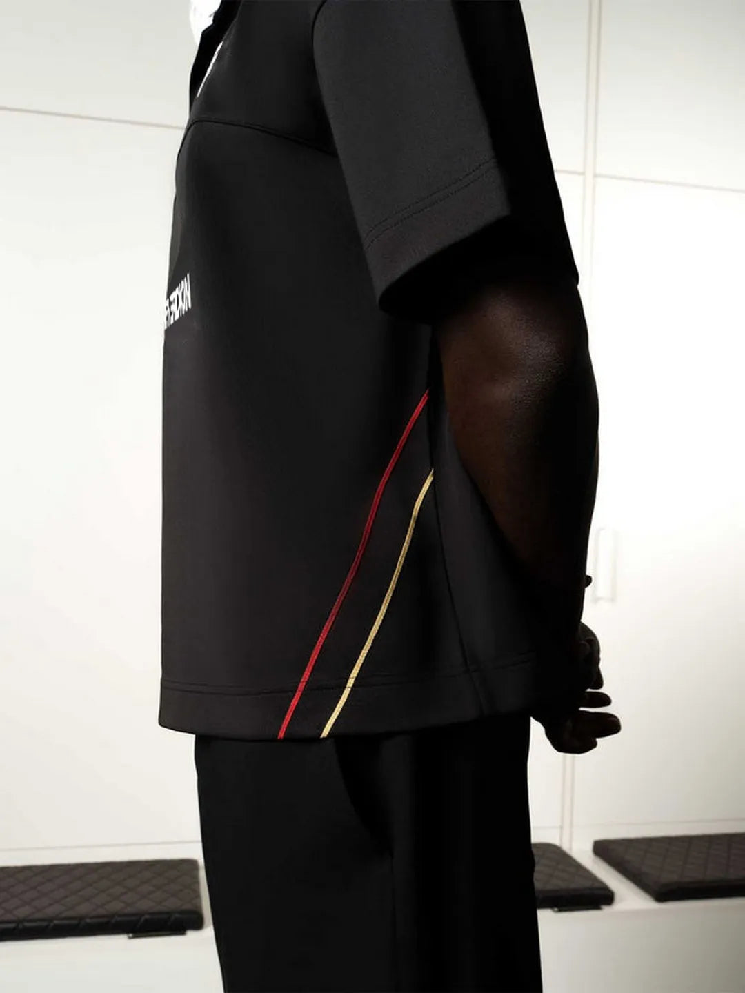 Person wearing a black outfit with red and yellow accents, standing against a white tiled wall.