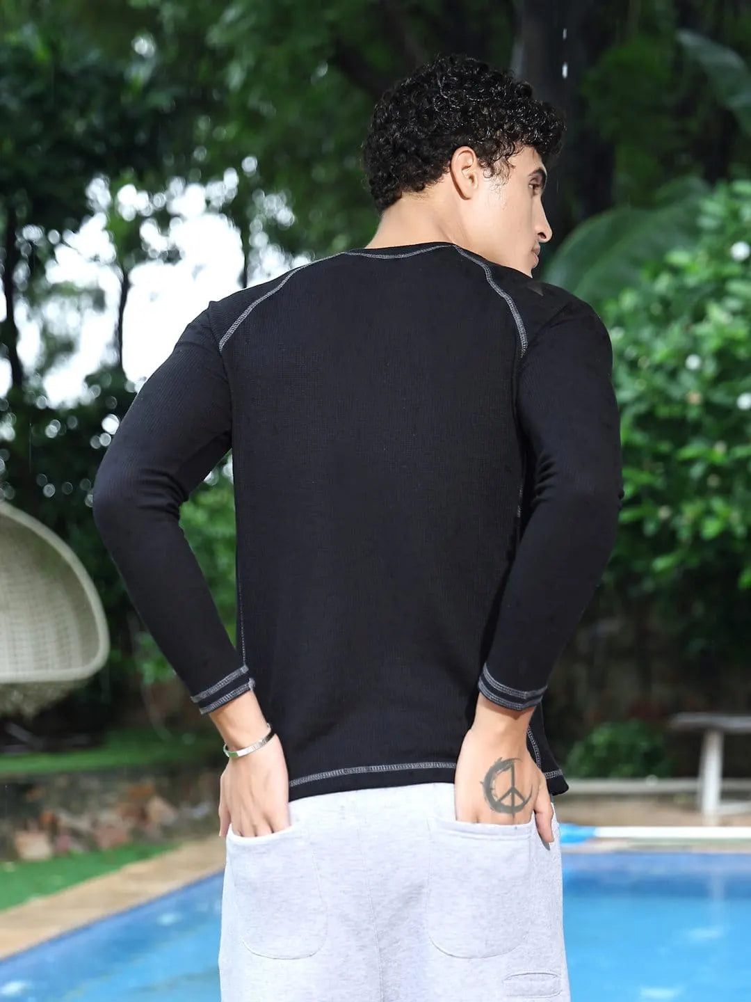 Person wearing a black long-sleeve shirt and white pants standing by a pool with greenery in the background