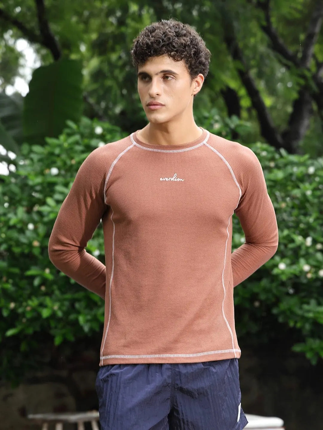 Man wearing a brown long-sleeve shirt with a brand logo, standing outdoors with greenery in the background.