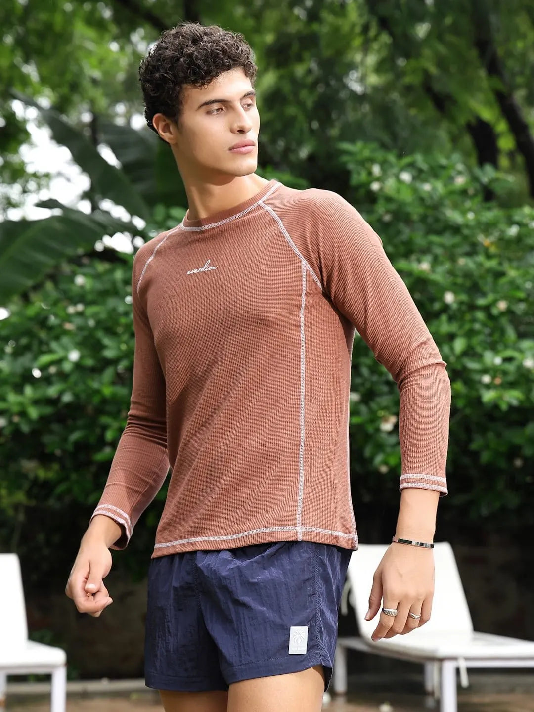 Man wearing a brown long-sleeve shirt and navy shorts outdoors with greenery in the background