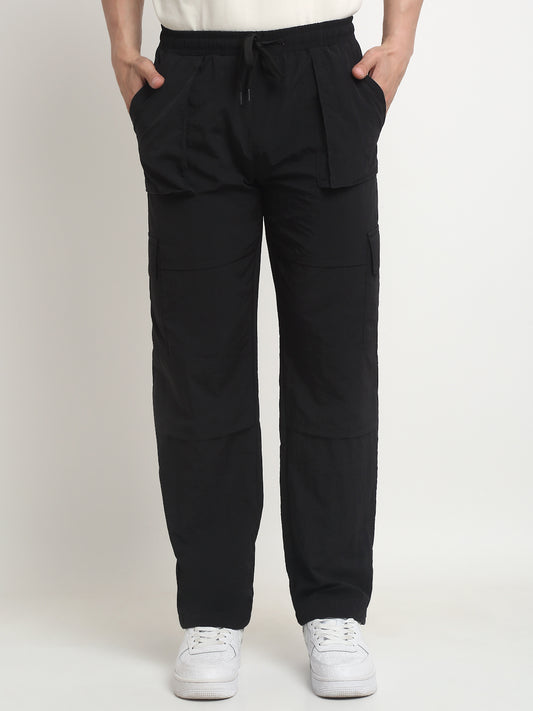 A person standing with hands in pockets, wearing black technical carpenter cargo pants and white sneakers.