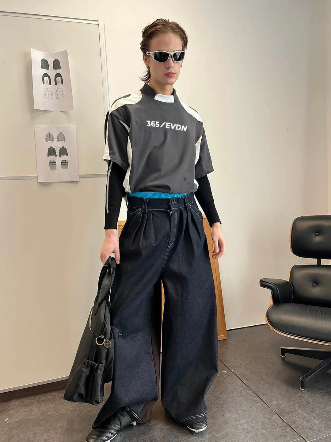 Person wearing a black and grey outfit with '365 EVDN' logo, holding a black bag in an indoor setting.