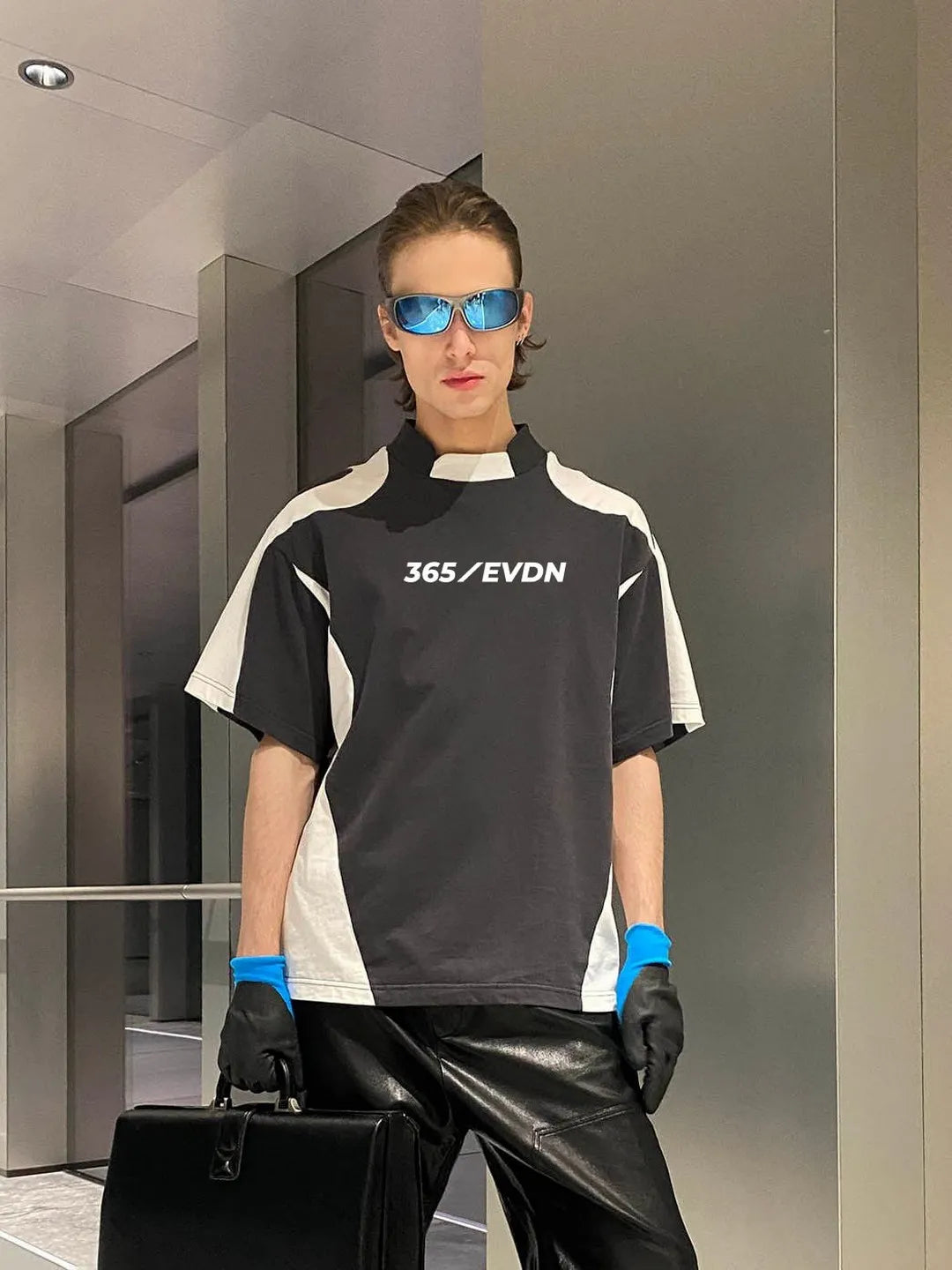 Person wearing a grey white shirt with '365/EVDN' text, black pants, and blue gloves, holding a briefcase.