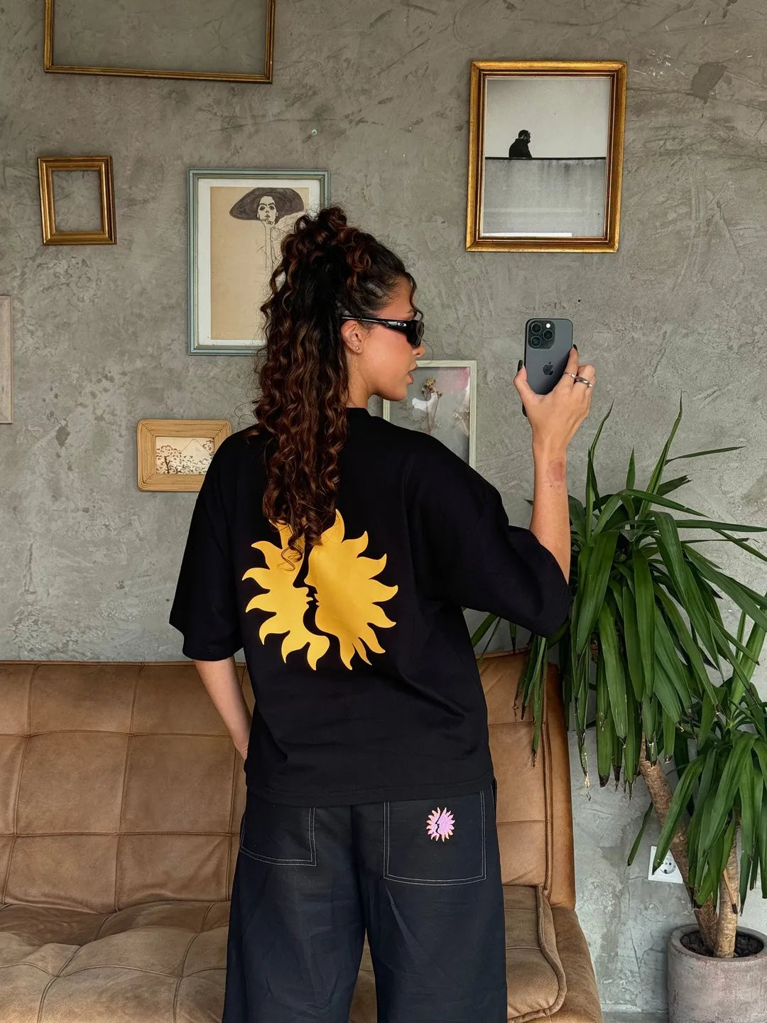 Person taking a photo of themselves in a mirror wearing a black t-shirt with a yellow sun design.