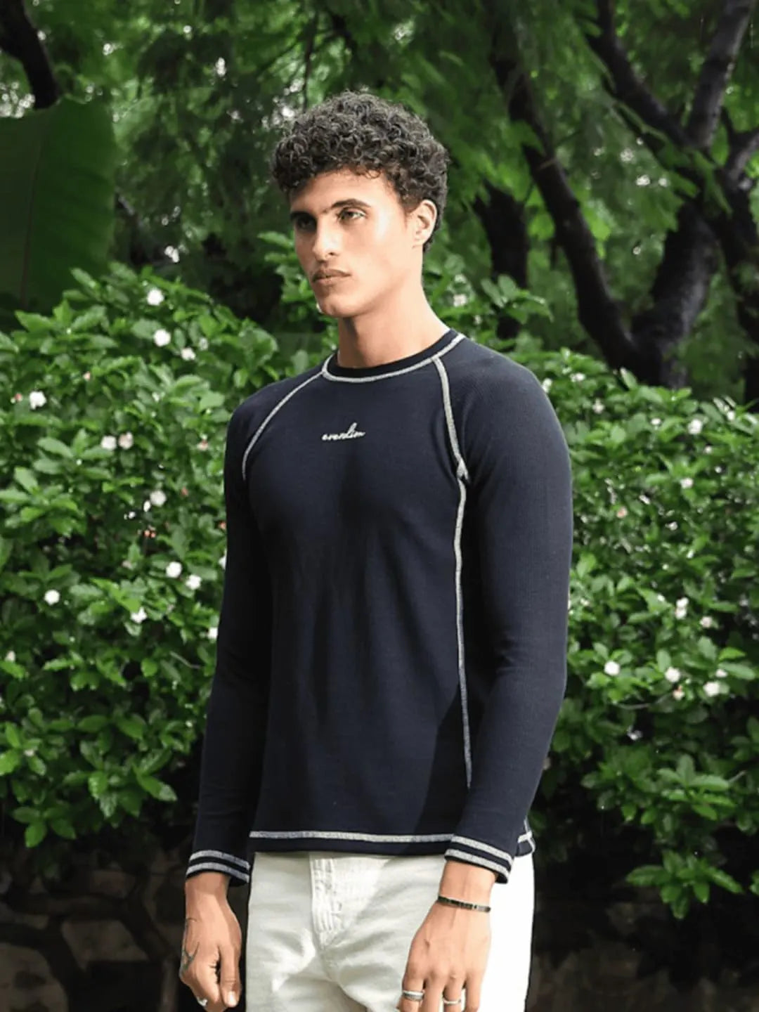 Man wearing a navy long-sleeve shirt with a brand logo, standing outdoors with greenery in the background