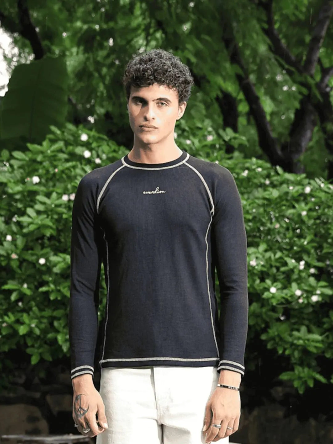 A man wearing a navy long-sleeved shirt with a brand logo stands outdoors, surrounded by greenery in the background.
