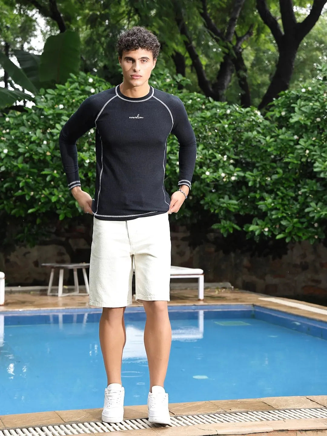 A man wearing a Navy long-sleeve shirt and white shorts standing by a pool with greenery in the background
