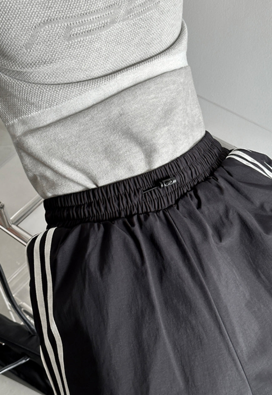 A person standing side-on, wearing striped parachute pants with contrast piping and an elasticated waistband with adjustable drawstrings.