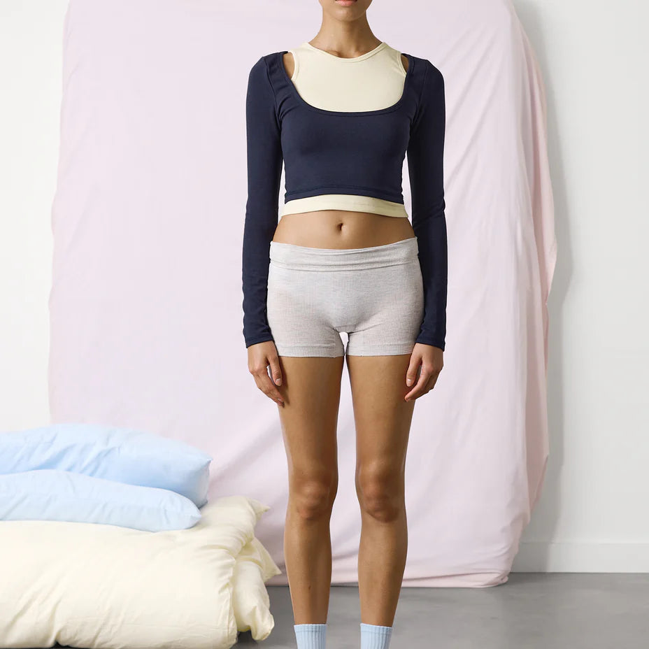 Person wearing a navy long-sleeve top and white shorts standing in a minimalistic room.