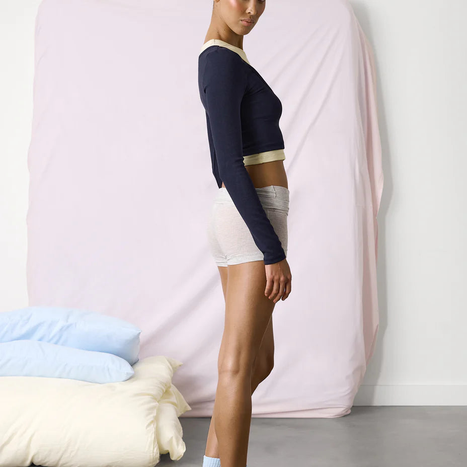 Woman wearing a navy long-sleeve top and white shorts standing against a pink curtain.