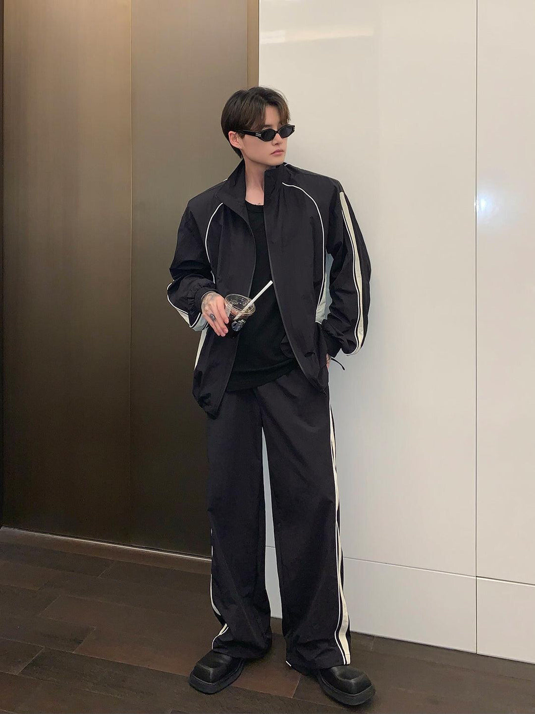 A man wearing a black tracksuit with white stripes on the pants, standing in front of a modern building.