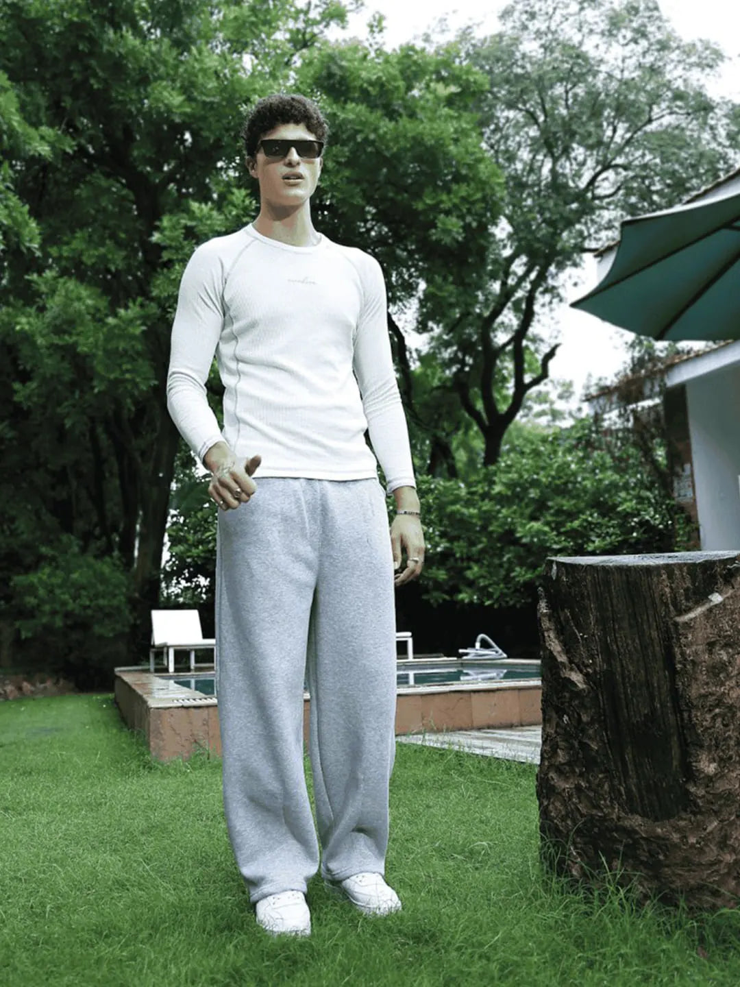 Person wearing a white long-sleeve shirt and gray sweatpants standing outdoors with trees and a pool in the background.