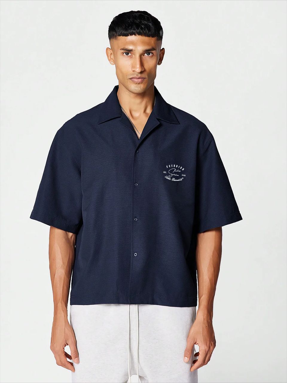 A man wearing a navy blue, oversized, half-sleeve bowling shirt with a crew neckline, featuring a small signature print on the front.
