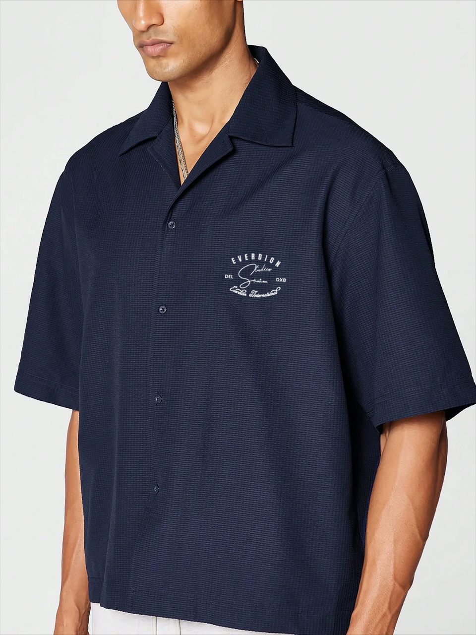 A man wearing a navy blue, oversized, half-sleeve bowling shirt with a crew neckline, featuring a small signature print on the front.