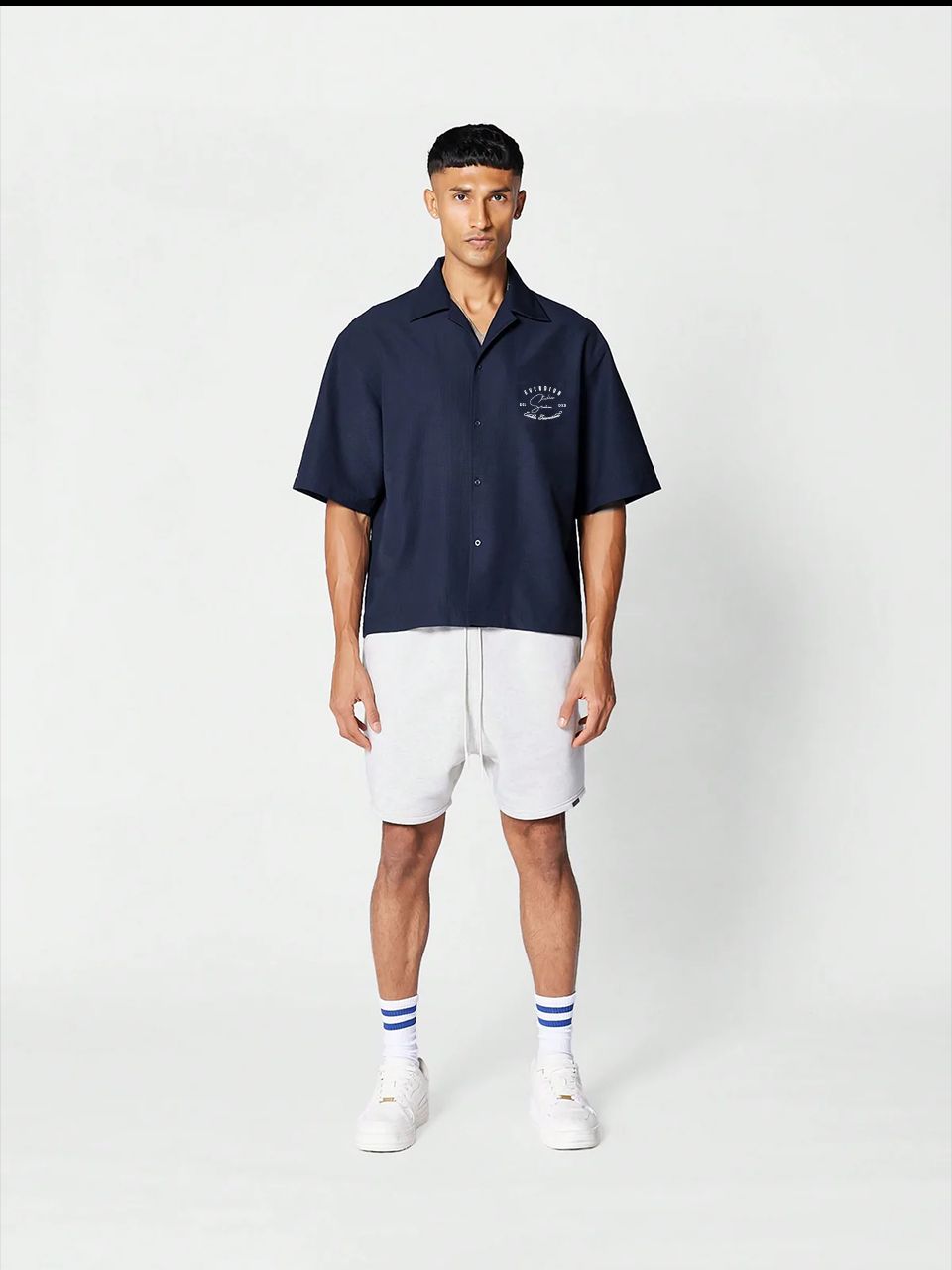 A man wearing a navy blue, oversized, half-sleeve bowling shirt with a crew neckline, featuring a small signature print on the front.