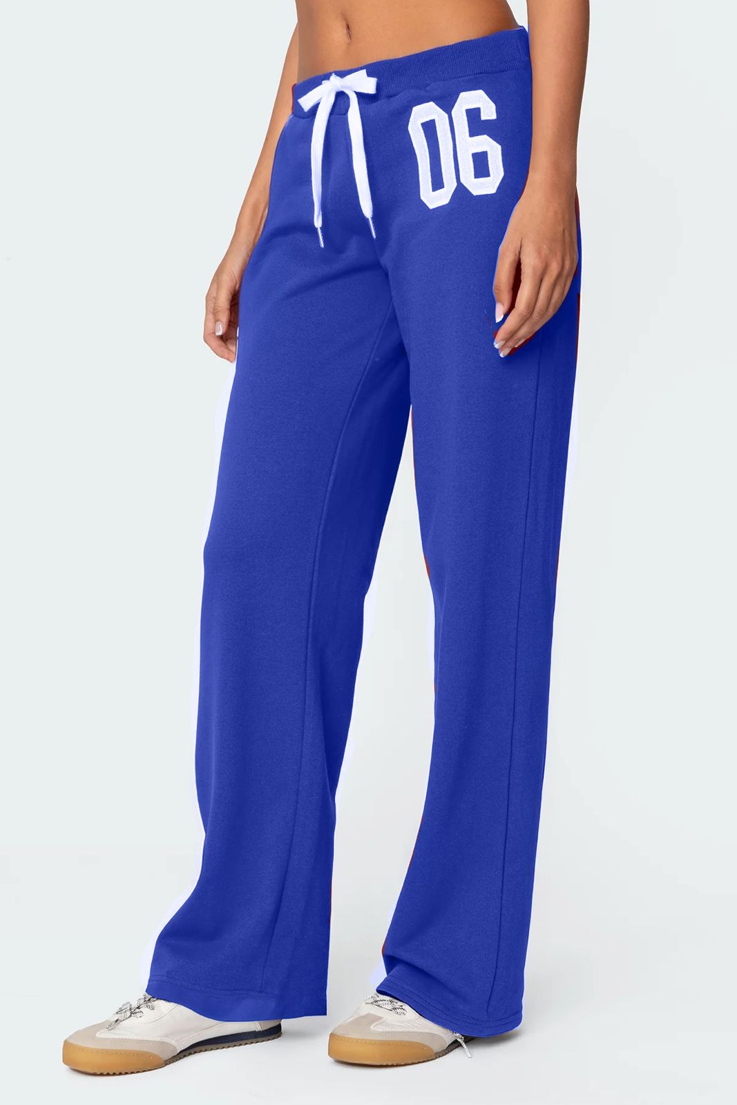A female model wearing a white tank top and blue lowrise trackpants with the number '06' printed on the left leg.