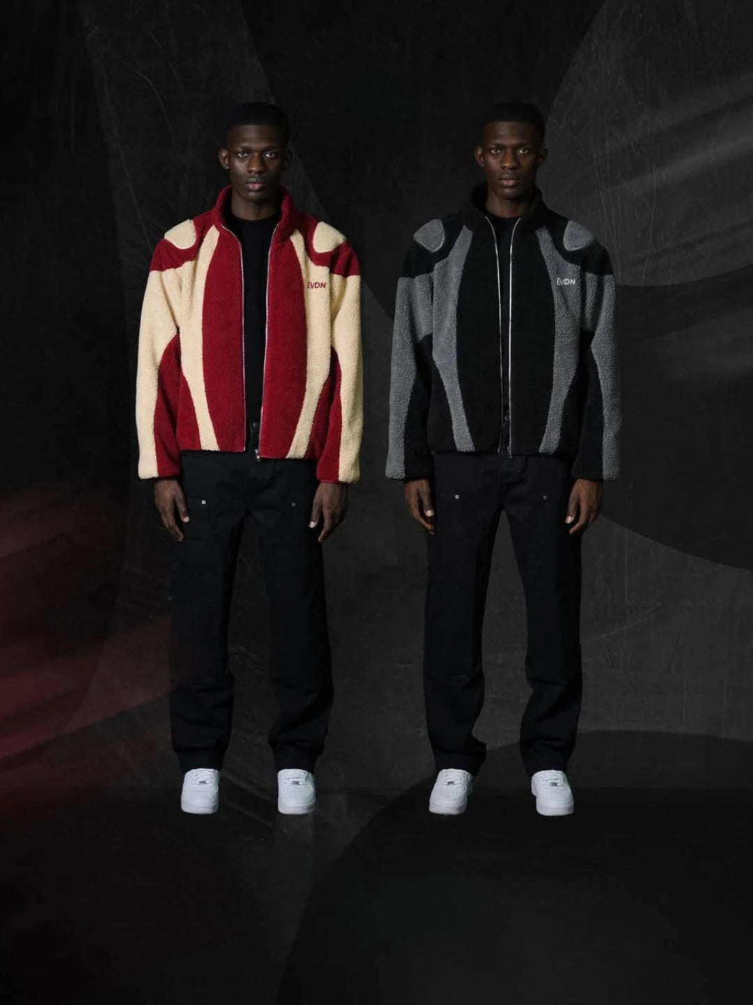 Two men wearing stylish jackets and pants standing against a dark background