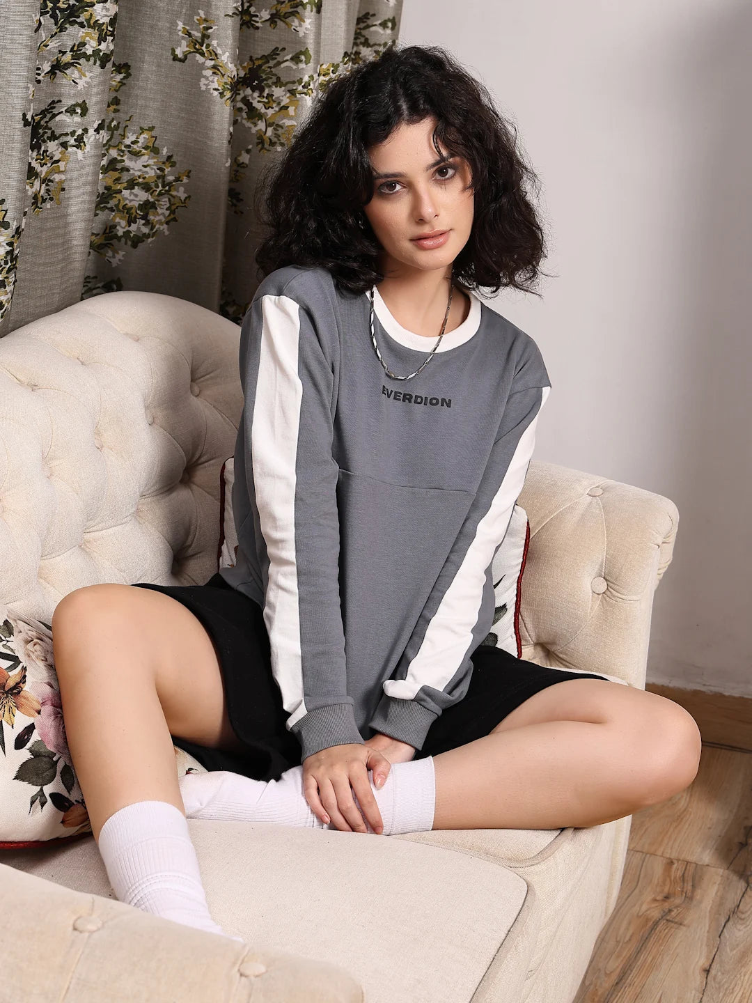Woman sitting on a beige armchair wearing a gray and white sweatshirt with text.