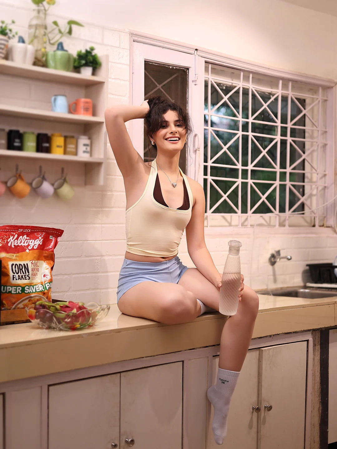 Woman sitting on a kitchen counter holding a bottle, with Kellogg's corn flakes and other snacks in the background.