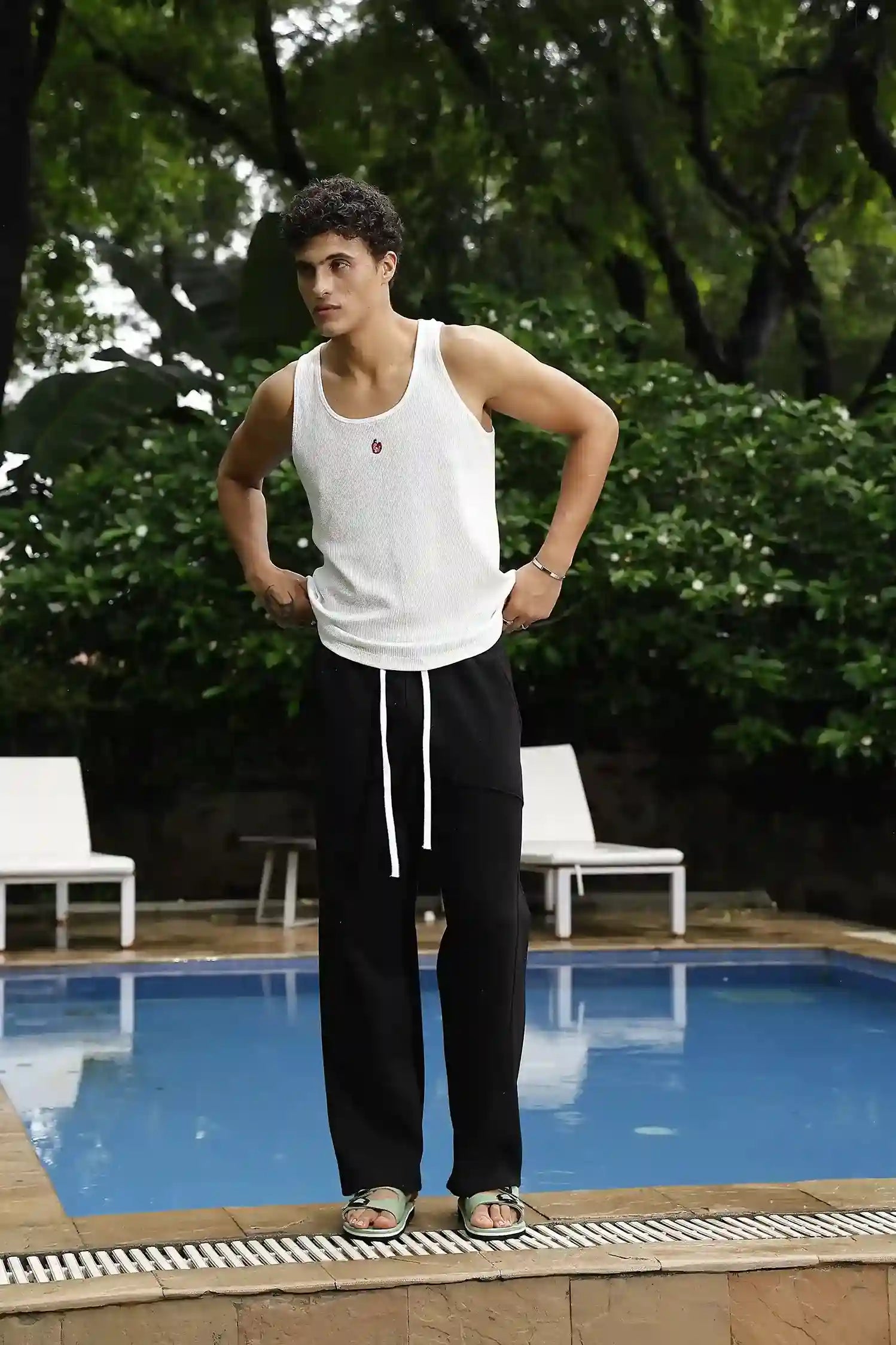 Person wearing a white tank top and black pants standing by a pool with greenery in the background