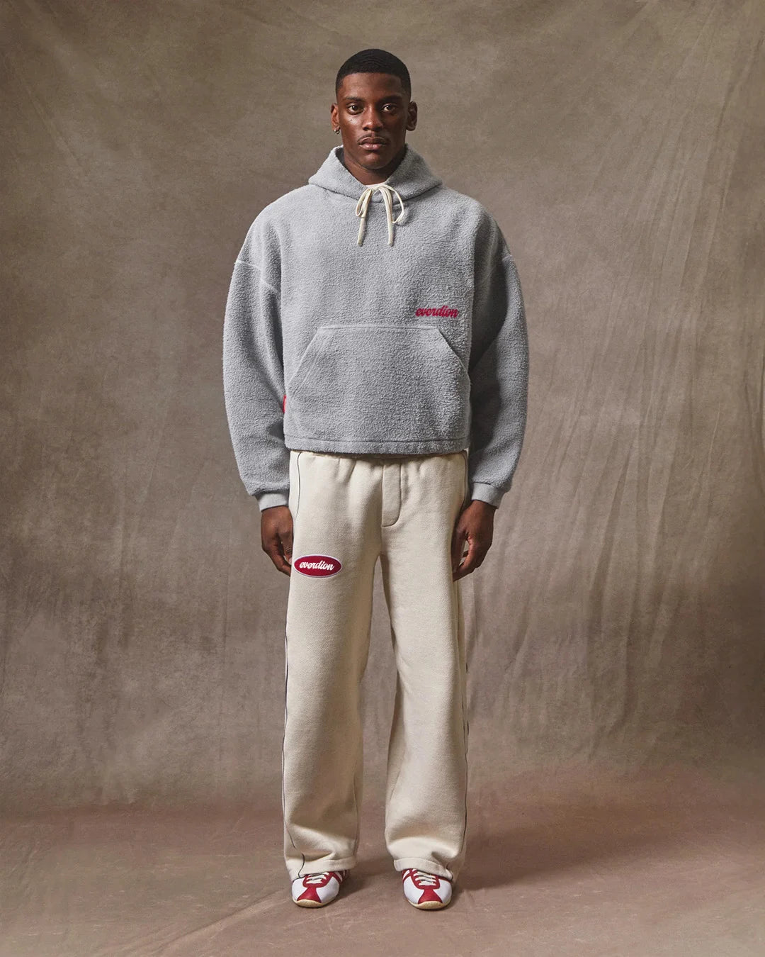 Person wearing a gray hoodie and beige pants with a logo, standing against a plain background