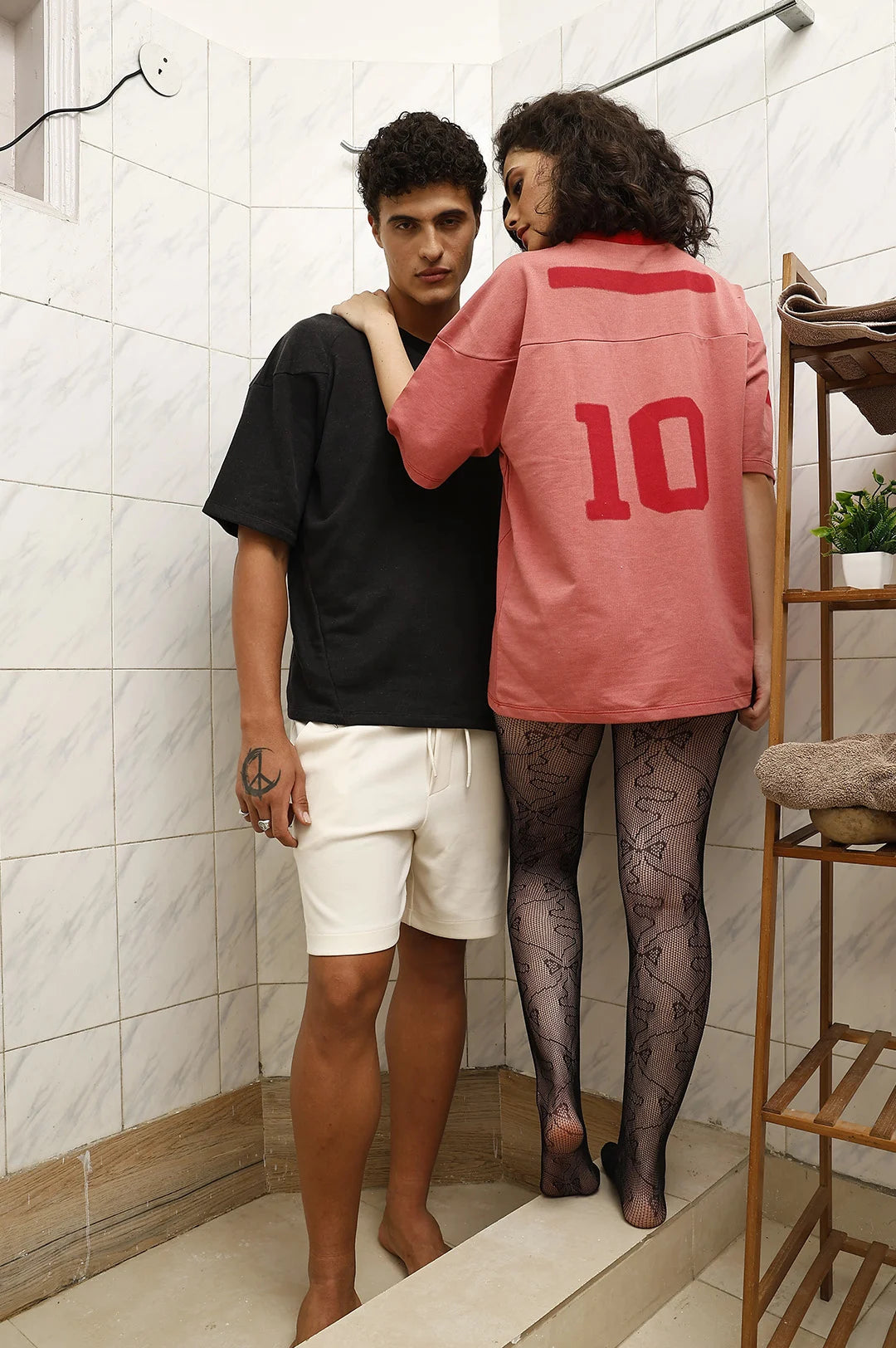 Two people standing in a bathroom, one wearing a black shirt and white shorts, the other in a pink shirt with the number 10.