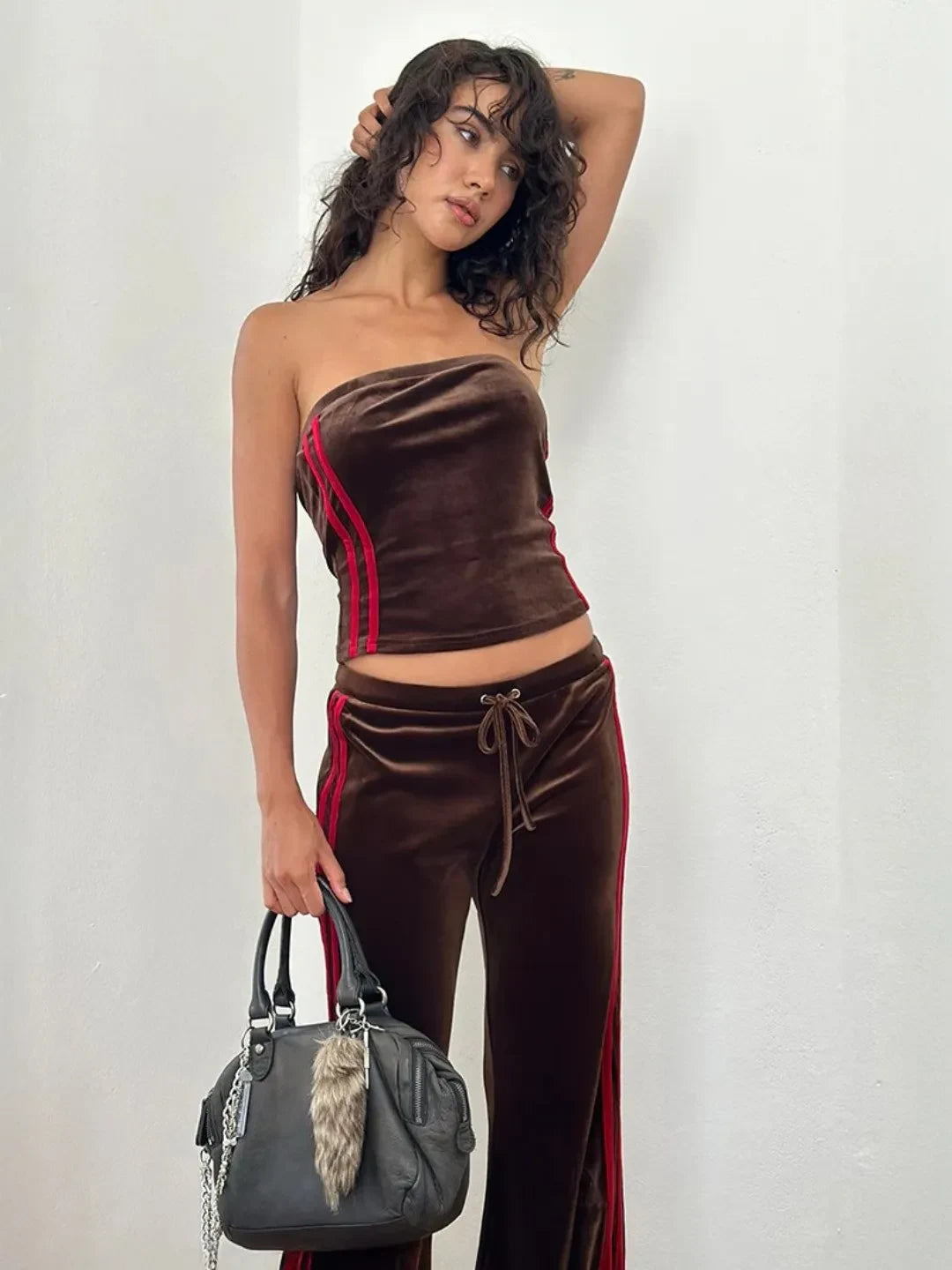 Woman wearing a brown velvet outfit with red accents, holding a gray handbag against a plain background.